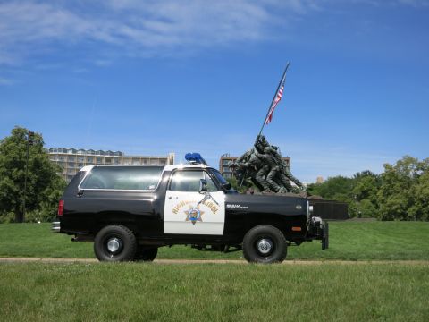 1991 Ford LTD Crown Victoria - Side view of a black-and-white Dodge Ram Charger Highway Patrol SUV parked on grass in front of a war memorial under a blue sky.