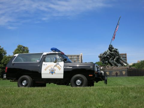 1991 Ford LTD Crown Victoria - black-and-white Dodge Ram Charger Highway Patrol SUV parked on a grassy field beside a bronze flag-raising memorial.