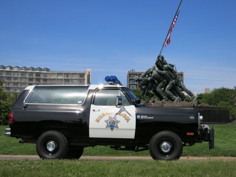 1991 Ford LTD Crown Victoria - black-and-white Dodge Ram Charger Highway Patrol SUV with rooftop light parked on grass before a flag-raising memorial.