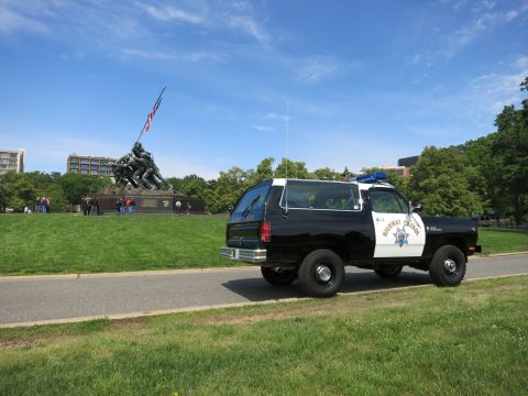 1991 Ford LTD Crown Victoria - black-and-white Highway Patrol SUV parked on a path with a flag-raising war memorial and onlookers in the background.