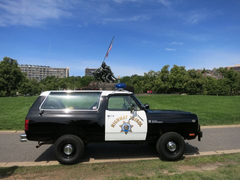 1991 Ford LTD Crown Victoria - Driver-side profile of a black-and-white Dodge Ram Charger Highway Patrol SUV parked on grass before a war memorial.
