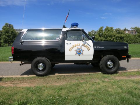 1991 Ford LTD Crown Victoria - Sunlit driver-side profile of a black-and-white Dodge Ram Charger Highway Patrol SUV parked on grass with trees behind.