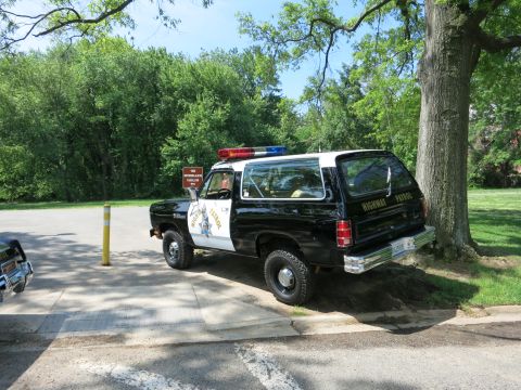 1991 Ford LTD Crown Victoria - black-and-white Dodge Ramcharger SUV marked Highway Patrol is parked beside a large tree with a smaller car at left.