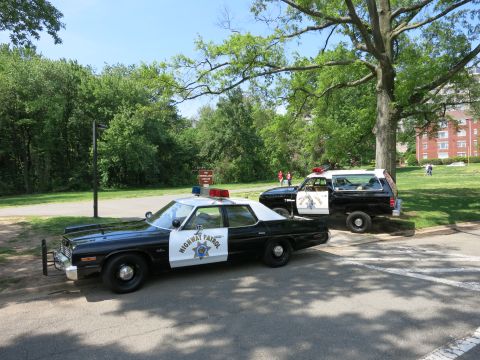 1991 Ford LTD Crown Victoria - black-and-white Highway Patrol sedan and a marked SUV are parked together in a park near trees and a brick building.