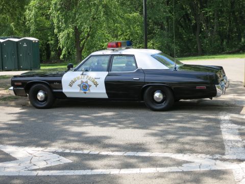 1991 Ford LTD Crown Victoria - black-and-white Highway Patrol sedan with a roof light bar is parked in a park beside green portable toilets.