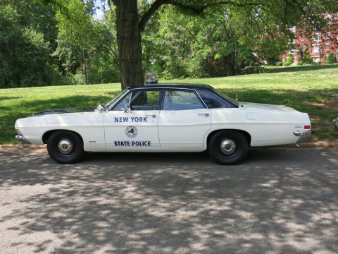 1991 Ford LTD Crown Victoria - white Ford four-door sedan marked New York State Police with a roof beacon is parked in front of grass and trees.
