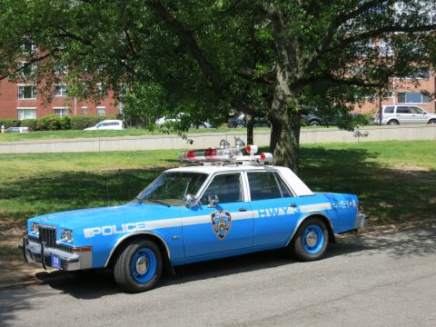 1991 Ford LTD Crown Victoria - blue-and-white NYPD police sedan with a roof light bar and blue wheels is parked on grass near a brick building.