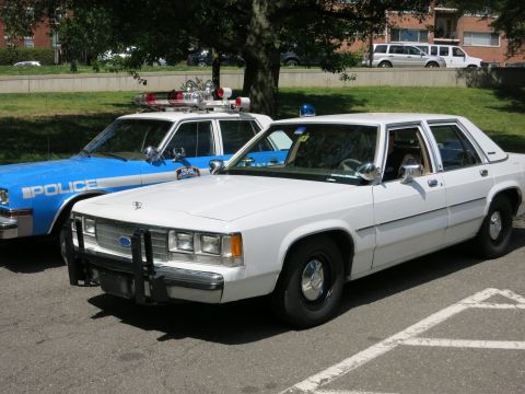 1991 Ford LTD Crown Victoria - white Ford police sedan with a brush guard is parked next to a blue NYPD car with a roof light bar.