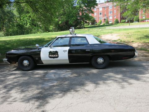 1991 Ford LTD Crown Victoria - black-and-white police four-door sedan with a roof light bar and door shield is parked in a shaded park.