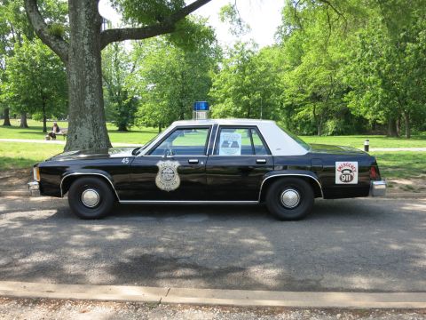 1991 Ford LTD Crown Victoria - black police four-door sedan numbered 45 with a door shield and a 911 decal sits parked under trees in a park.