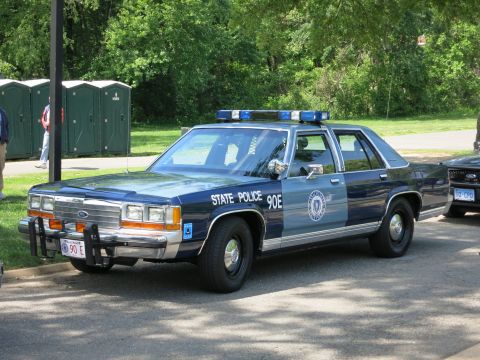 1991 Ford LTD Crown Victoria - dark-blue Ford State Police sedan marked 90E with a brush guard and roof lights is parked near portable toilets.