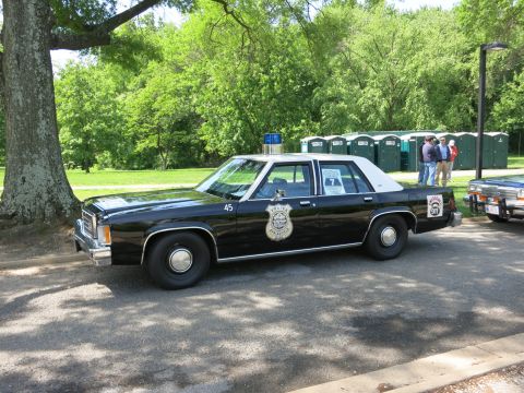 1991 Ford LTD Crown Victoria - black police four-door sedan numbered 45 with a door shield and roof beacon is parked under trees near a row of toilets.