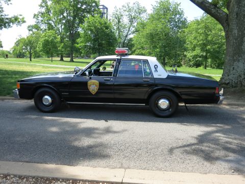 1991 Ford LTD Crown Victoria - black-and-white police sedan with a white roof, roof light bar and door shield is parked along a park road.