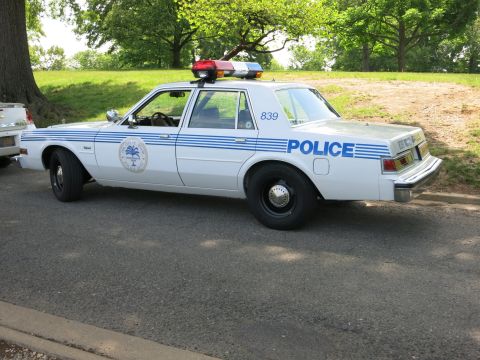 1991 Ford LTD Crown Victoria - white police sedan with blue striping, unit number 839, a door agency seal and a red-and-blue roof light bar parked on a road.