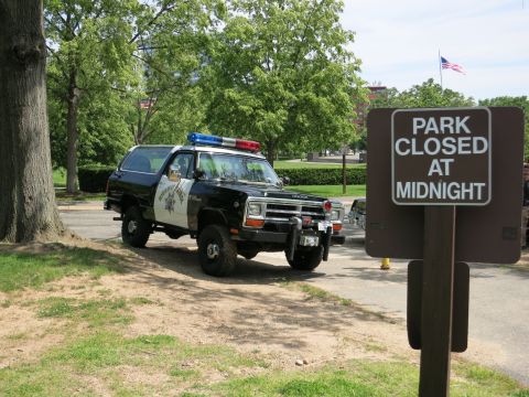 1991 Ford LTD Crown Victoria - black-and-white Dodge Ramcharger SUV in police livery with a roof light bar parked beyond a brown 'PARK CLOSED AT MIDNIGHT' sign.