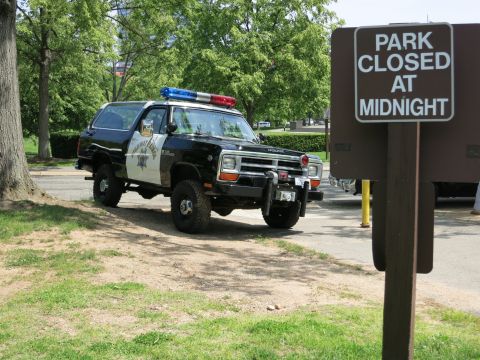 1991 Ford LTD Crown Victoria - black-and-white Dodge Ramcharger police SUV with a roof light bar and front winch parked next to a 'PARK CLOSED AT MIDNIGHT' sign.