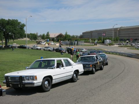 1991 Ford LTD Crown Victoria - line of police cars, led by a white sedan with a push bumper, parked along a road with the Pentagon building in the background.