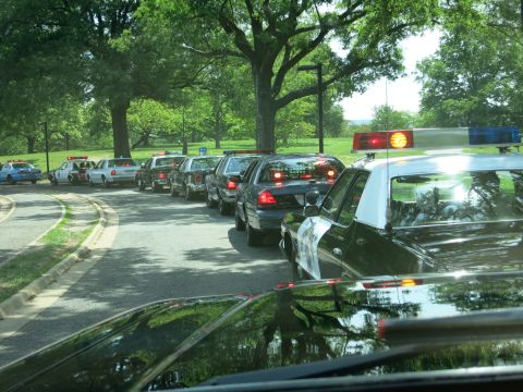 1991 Ford LTD Crown Victoria - queue of police cars with illuminated brake and roof lights waits along a shaded road, viewed over the hood of a lead car.