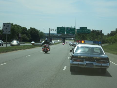1991 Ford LTD Crown Victoria - black 'HIGHWAY PATROL' sedan and a flag-bearing motorcycle travel a highway under green signs for Alexandria and Washington.
