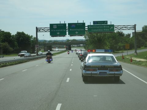 1991 Ford LTD Crown Victoria - convoy of police vehicles led by a motorcycle drives a highway under green signs reading Alexandria, Washington and Columbia Pike.