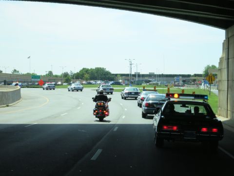 1991 Ford LTD Crown Victoria - line of police cars and a motorcycle emerge from a highway underpass onto a curving ramp with brake lights glowing.