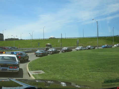 1991 Ford LTD Crown Victoria - Several police cars are staged along a roadway below a grassy embankment and overpass, viewed from inside a lead vehicle.