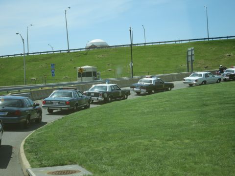 1991 Ford LTD Crown Victoria - mix of police sedans, including a black-and-white unit, parked along a roadway beside a grassy embankment under a clear sky.