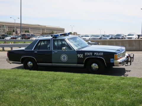 1991 Ford LTD Crown Victoria - dark-blue State Police sedan marked 90E with a roof lightbar and push bumper sits on grass in front of the Pentagon.