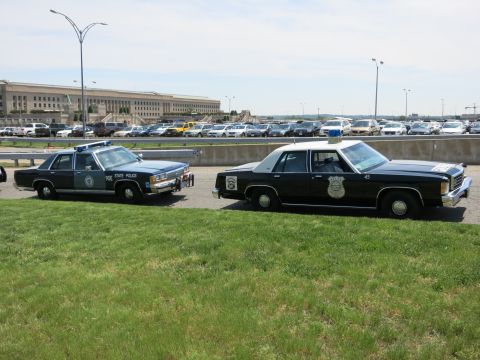 1991 Ford LTD Crown Victoria - Two dark vintage police sedans, including a State Police cruiser, are parked side by side before the Pentagon.