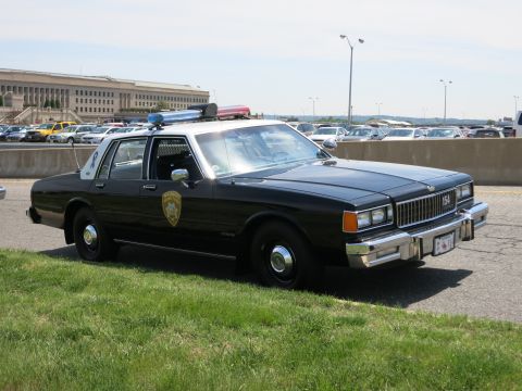 1991 Ford LTD Crown Victoria - black police sedan numbered 154 with a roof lightbar and door seal decal is parked on grass before the Pentagon.