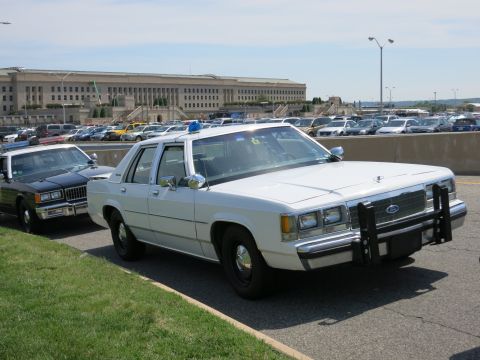 1991 Ford LTD Crown Victoria - white Ford police sedan with a roof lightbar and chrome push bumper is parked beside another car near the Pentagon.