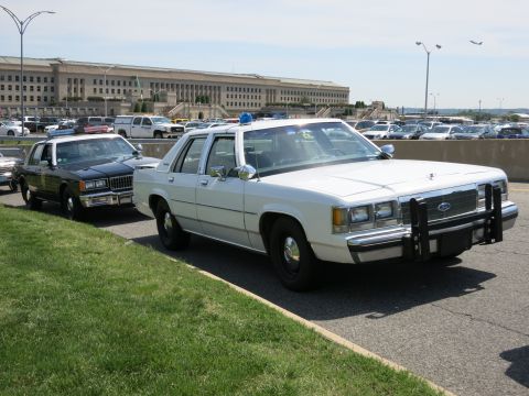 1991 Ford LTD Crown Victoria - white Ford police sedan with roof lights and a push bumper is parked at an angle on grass before the Pentagon.