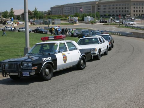1991 Ford LTD Crown Victoria - row of black-and-white Police sedans is lined up along a road, with people gathered near the Pentagon grounds.