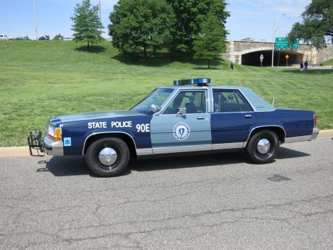 1991 Ford LTD Crown Victoria - two-tone blue State Police sedan marked 90E with a state seal decal is parked on a road beside a grassy hillside.