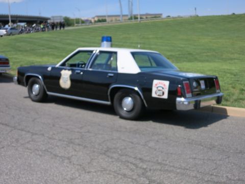 1991 Ford LTD Crown Victoria - Rear three-quarter view of a black-and-white police sedan with a gold badge door decal parked near a grassy hill.