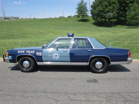 1991 Ford LTD Crown Victoria - Profile of a two-tone blue State Police sedan marked 90E with door seal decal parked on a road by a grassy slope.