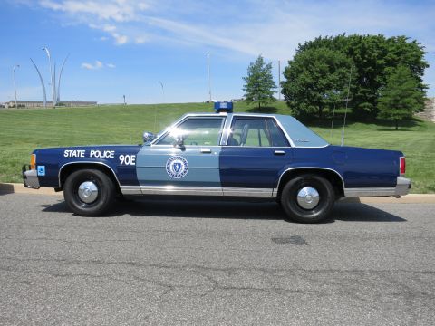 1991 Ford LTD Crown Victoria - two-tone blue State Police sedan marked 90E with rear antennas is parked on a road beside a grassy hillside.