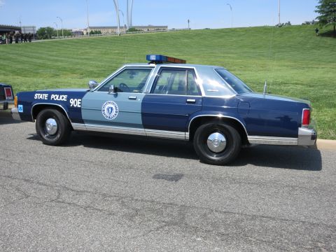 1991 Ford LTD Crown Victoria - Rear-quarter view of a two-tone blue State Police sedan marked 90E parked on a road in front of a grassy hill.