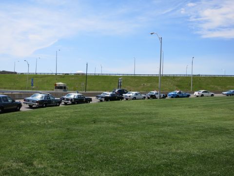 1991 Ford LTD Crown Victoria - distant row of cars and a small bus parked along a roadway beside grassy embankments and an overpass.