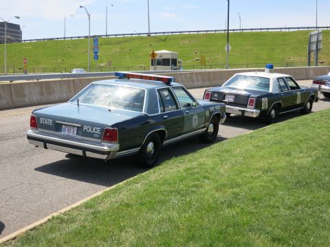 1991 Ford LTD Crown Victoria - two-tone blue STATE POLICE sedan parked at a curb in front of a tan vintage sedan along a roadway.