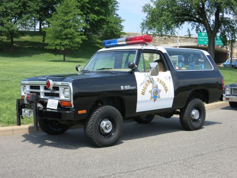 1991 Ford LTD Crown Victoria - black-and-white Dodge Ram Charger Highway Patrol SUV with roof light bar parked on a road near an overpass.