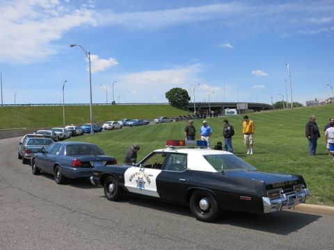 1991 Ford LTD Crown Victoria - black-and-white Highway Patrol sedan parked at the head of a long line of cars beside a grassy embankment.
