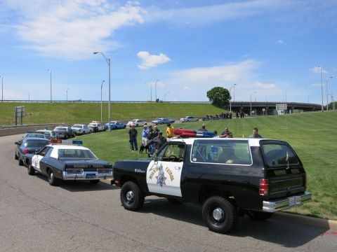 1991 Ford LTD Crown Victoria - black-and-white Dodge Highway Patrol SUV and sedan parked near a grassy hill as a crowd gathers behind them.
