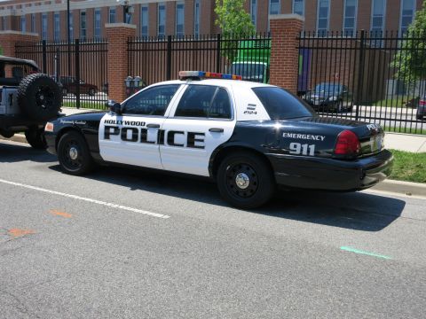 1991 Ford LTD Crown Victoria - black-and-white Hollywood Police Ford Crown Victoria marked EMERGENCY 911 parked at a curb by a fence.