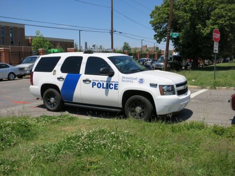1991 Ford LTD Crown Victoria - white Federal Protective Service Police Chevrolet Tahoe SUV marked Homeland Security parked amid tall grass.
