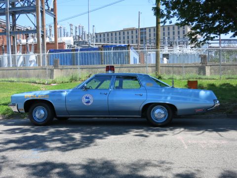 1991 Ford LTD Crown Victoria - vintage light-blue full-size police sedan with a red roof light marked POLICE parked beside a fenced industrial yard.