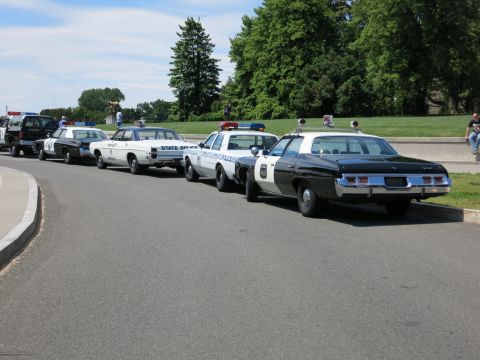 1991 Ford LTD Crown Victoria - line of vintage police cruisers including a black-and-white and a State Police car parked along a curved roadway.