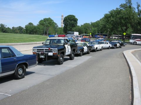 1991 Ford LTD Crown Victoria - line of police vehicles led by a black-and-white Dodge SUV parked along a road with the Washington Monument behind.