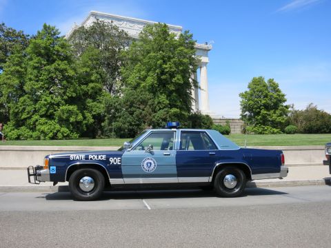1991 Ford LTD Crown Victoria - blue-and-silver Massachusetts State Police Ford Crown Victoria marked STATE POLICE 90E parked before a stone memorial.