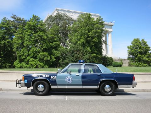 1991 Ford LTD Crown Victoria - two-tone blue State Police sedan marked '90E' sits curbside in front of a white columned marble building.